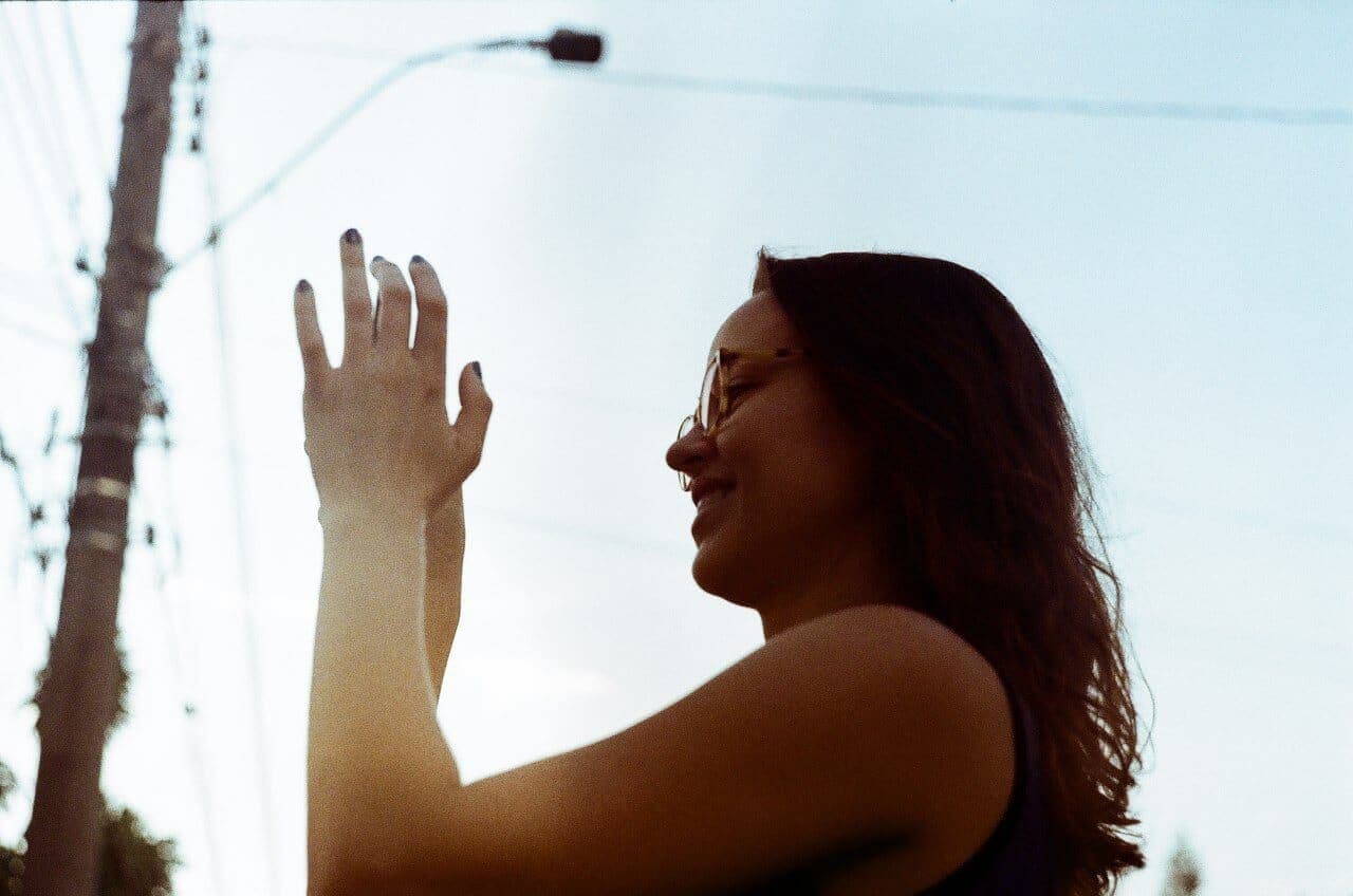 A foto mostra Prussiana contra a luz, tirando uma foto. Ela é uma mulher branca, usando óculos, de cabelos compridos. Olha para a esquerda e sorri.