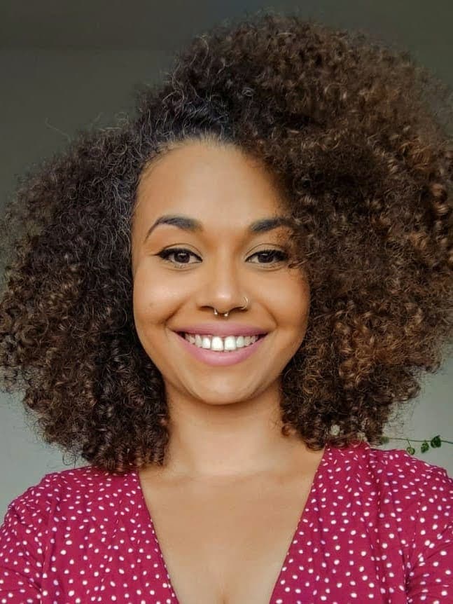 Amanda é uma mulher negra de pele clara, e na foto está com uma roupa avermelhada, com bolinhas brancas. Ela está virada de frente e olha diretamente para a câmera. Amanda está sorrindo, possui cabelos cacheados black power na altura dos ombros, olhos castanhos e tem piercing no nariz.