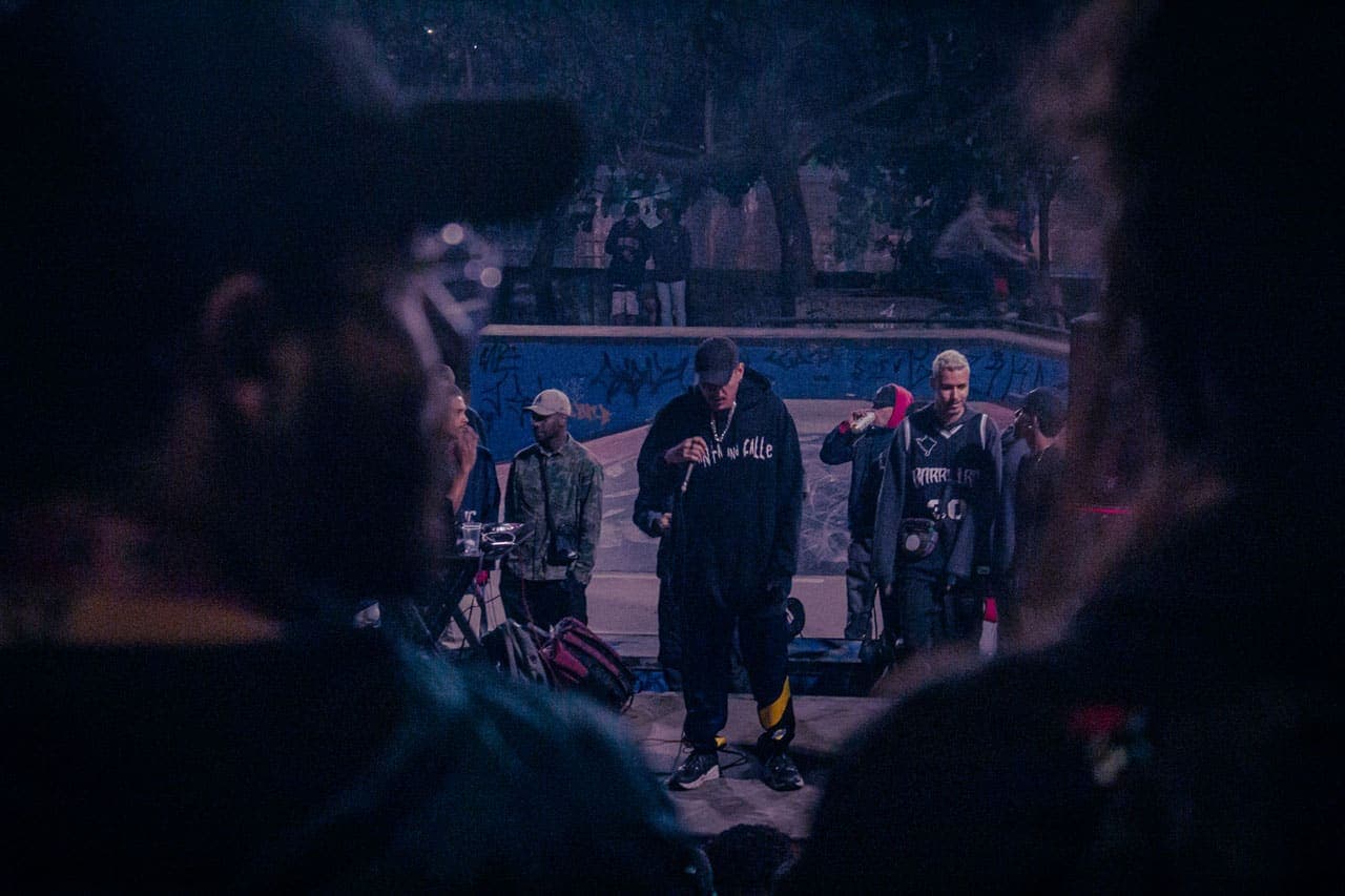 A imagem traz, em um primeiro plano, a silhueta de duas pessoas que assistem, das arquibancadas da Praça Cristo Reina, à apresentação de um grupo de jovens que, ao fundo, participam de uma batalha de MCs na Pista de Skate do Barreiro.