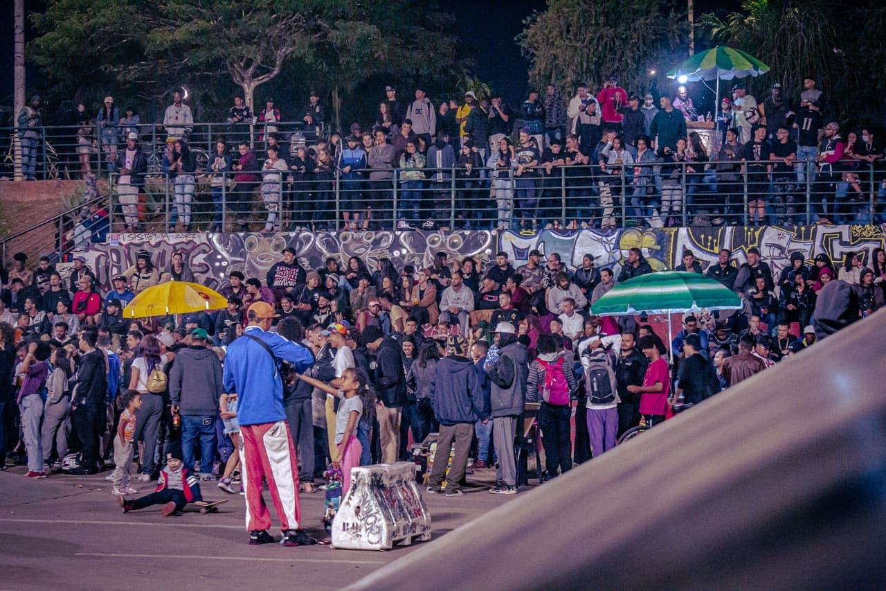 A imagem retrata uma arquibancada tomada por um grande número de jovens que assistem ao evento FaráOeste, na Praça Cristo Reina, localizada no Barreiro. Em meio ao público, despontam os guarda-sóis dos ambulantes que vendem bebidas. Em um primeiro plano, crianças brincam com seus skates.