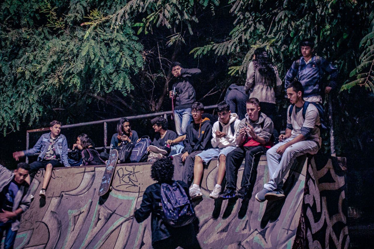  A imagem traz um grupo de adolescentes que, portando suas mochilas, sentam-se para conversar no alto da estrutura de uma pista de skate do tipo “half pipe”.