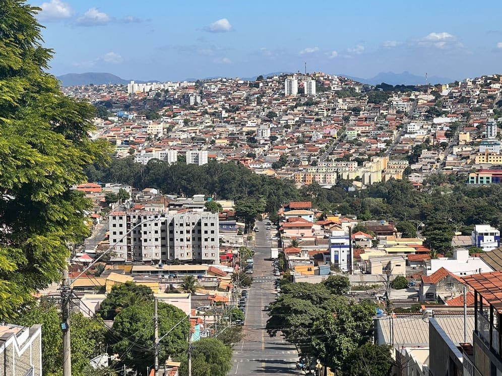 A imagem captura uma paisagem urbana do Barreiro. Ao centro, uma rua se estende até ir de encontro às árvores que cortam o cenário, em um segundo plano. Ao fundo, em um aclive, edificações diversas compõem uma área densamente povoada.