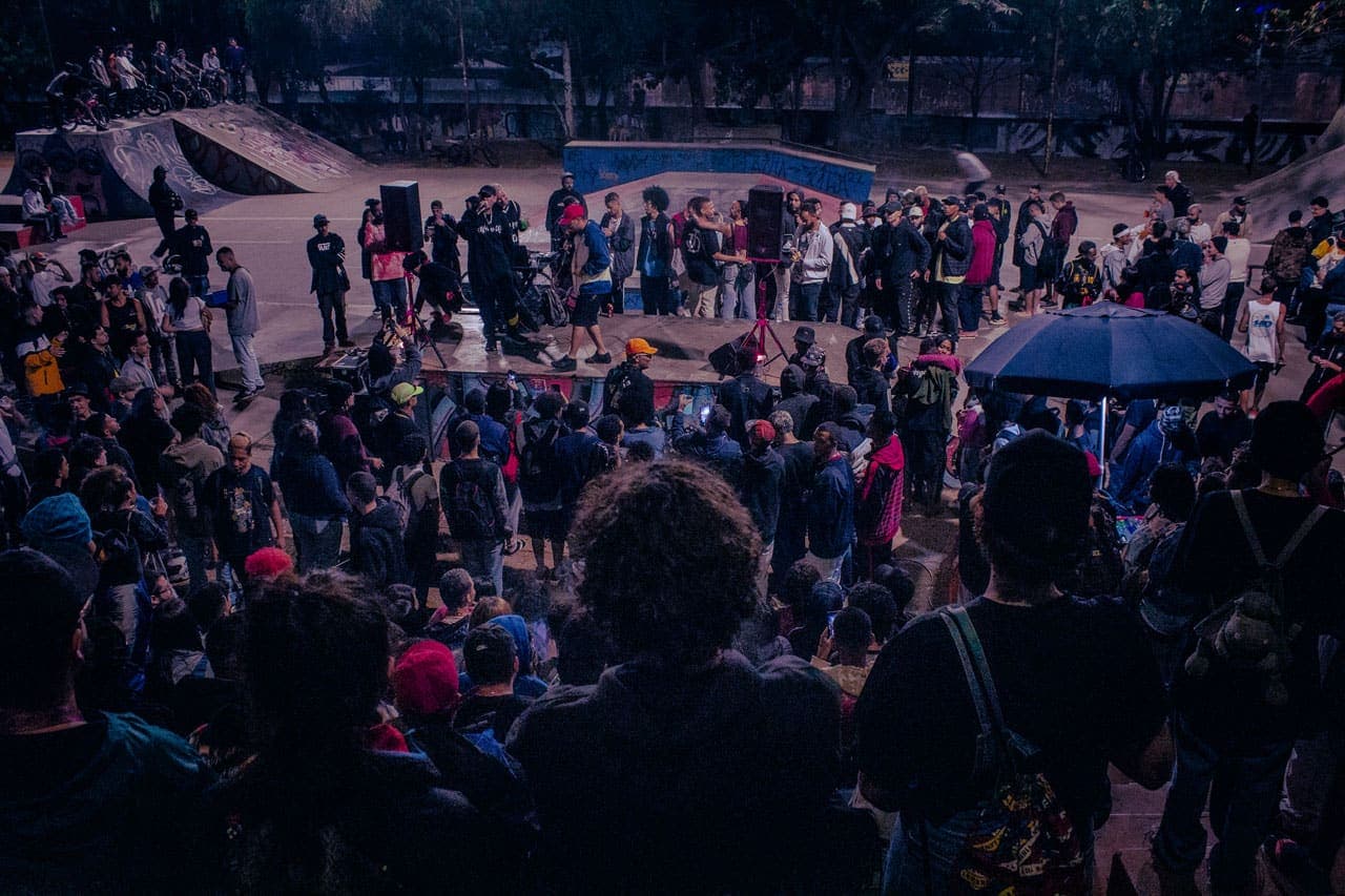 A imagem traz uma multidão de jovens que ocupam as arquibancadas e um palco montado na Pista de Skate do Barreiro. Todos os olhares estão voltados para as duas figuras que, ao centro, travam entre si uma batalha de MCs no tradicional evento FaráOeste.