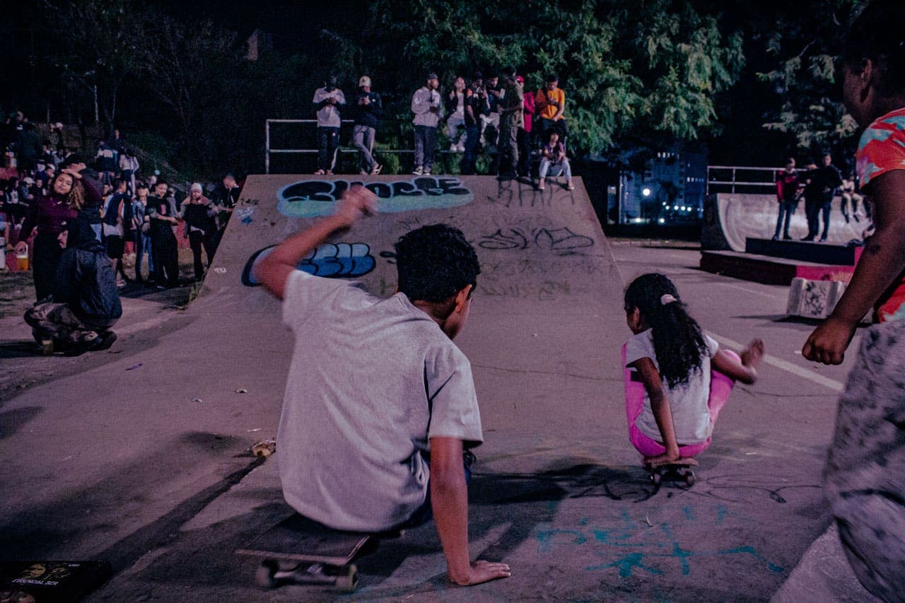 A imagem traz, em um primeiro plano, dois jovens – um rapaz adolescente e uma pequena garota – que, sentados em seus skates, usam as mãos para “remar” ao longo da pista. Ao fundo, um grande número de jovens assiste à batalha de MCs.