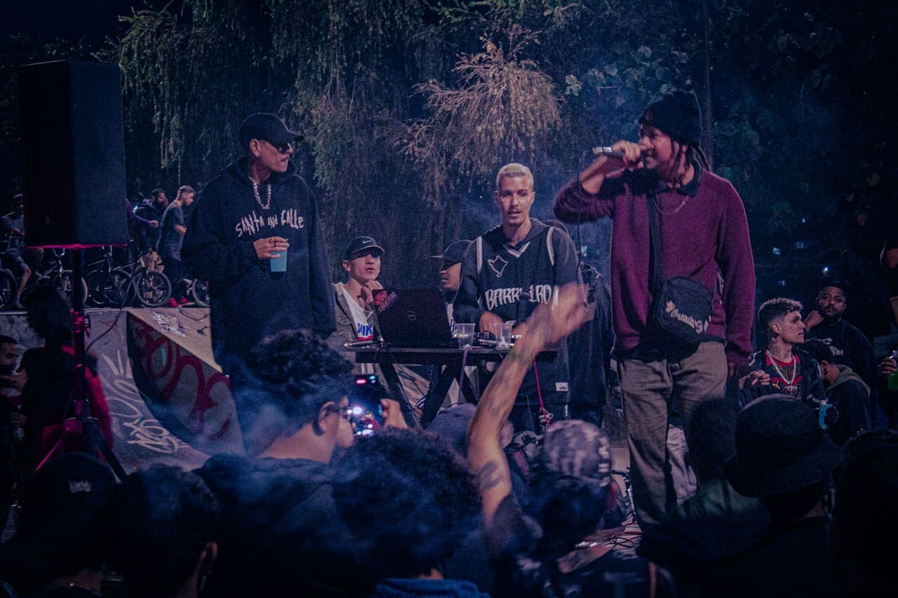 A imagem retrata, ao centro, três homens jovens que se apresentam no palco da Pista de Skate do Barreiro – dois MCs e um DJ. Suas vestes trazem estampados os dizeres “SANTA AND CALLE” e “BARREIRO”. Ao redor das três figuras, um grande número de pessoas, também jovens, acompanha o evento.