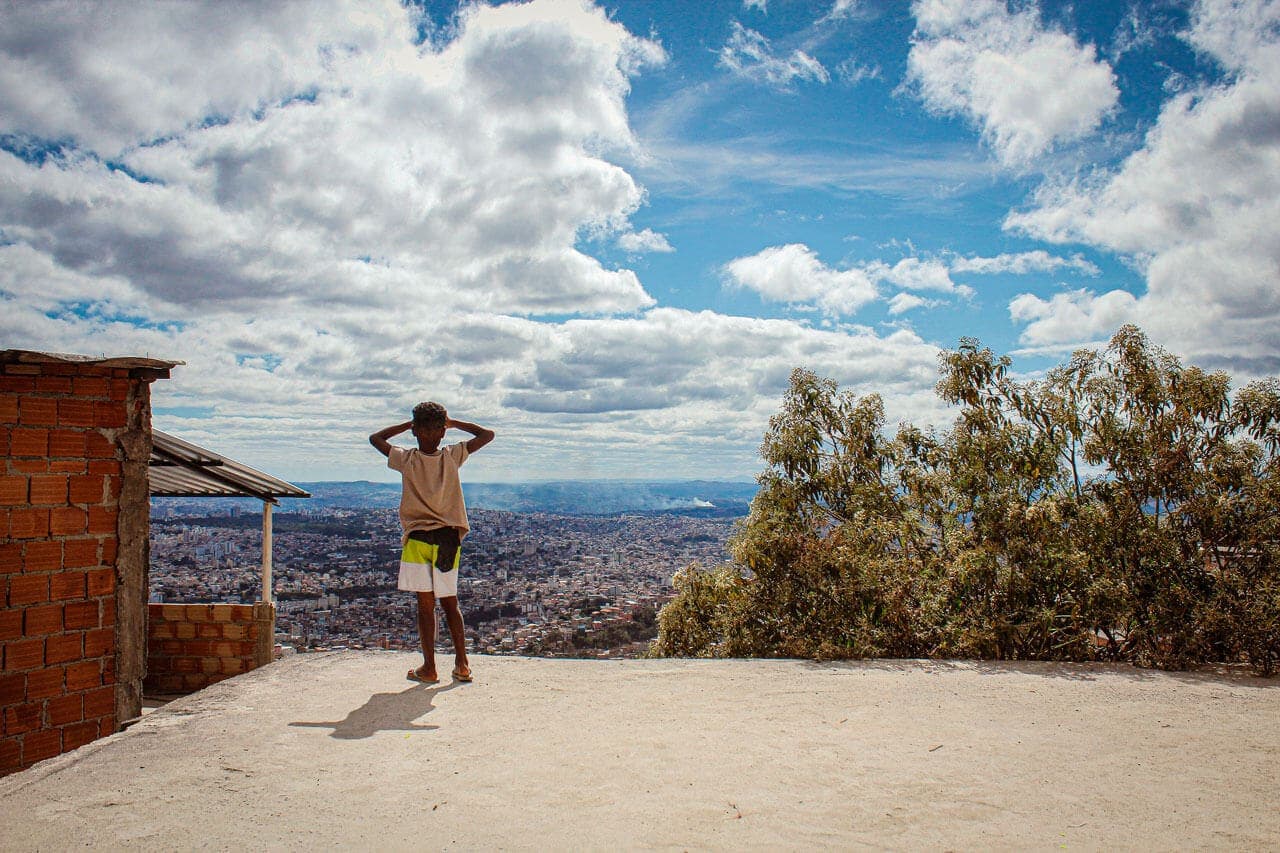 A imagem mostra uma criança de costas sobre uma laje em primeiro plano. O menino está com as mãos no rosto, protegendo os olhos do sol e observando a cidade, numa vista panorâmica.