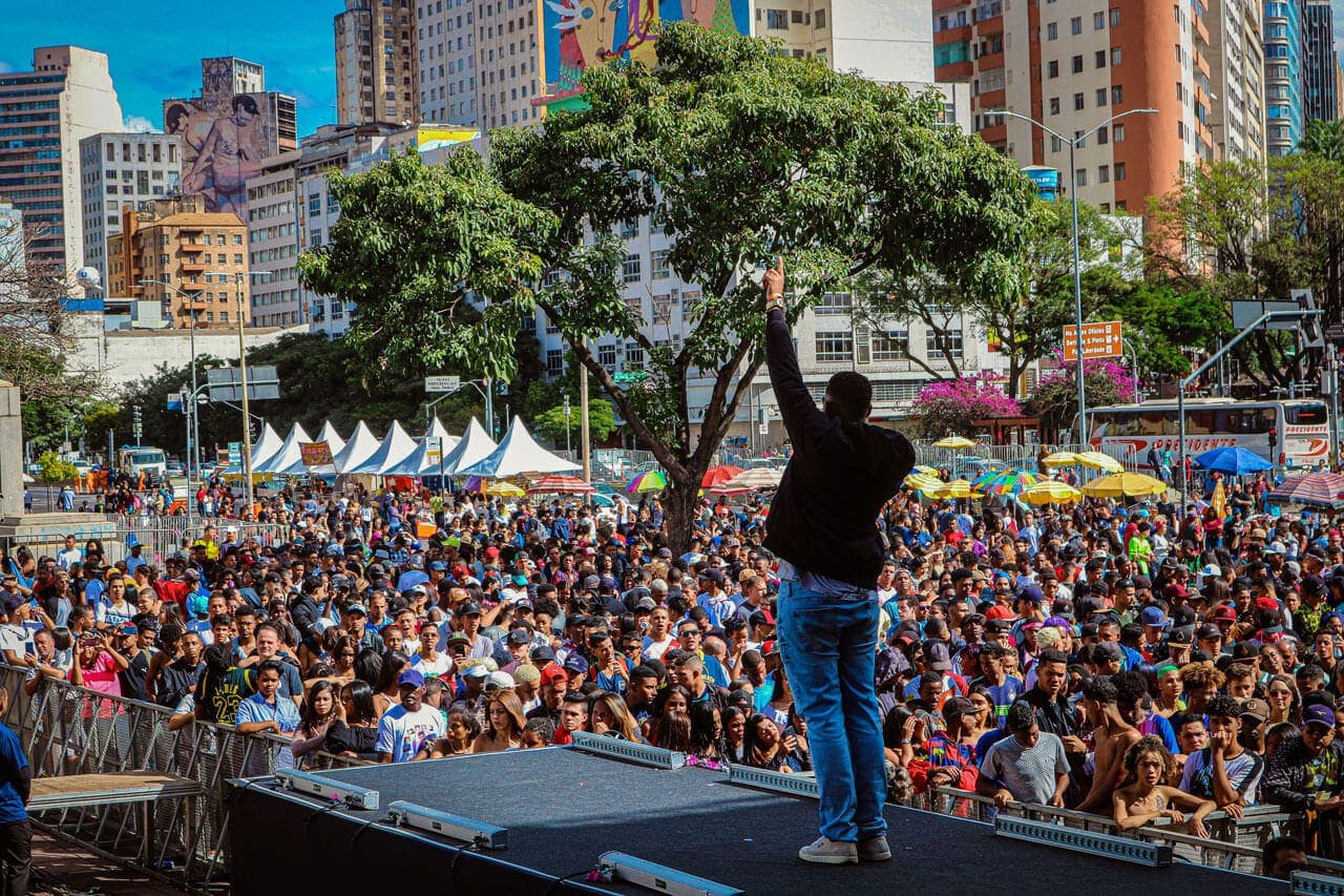 A imagem retrata o DJ Marcelo Mattos sobre o palco, de costas, em primeiro plano. Marcelo é um homem negro, e está vestido na foto com uma calça jeans, uma camisa de manga comprida na cor preta e tênis na cor bege. Ele está falando ao microfone, segurando com a mão direita, e o braço esquerdo está levantado apontando para cima. A sua frente está uma multidão, na Praça da Estação. É possível ver alguns guarda-sóis e barracas de apoio ao evento. Ao fundo é possível ver as edificações do entorno da Praça da Estação.