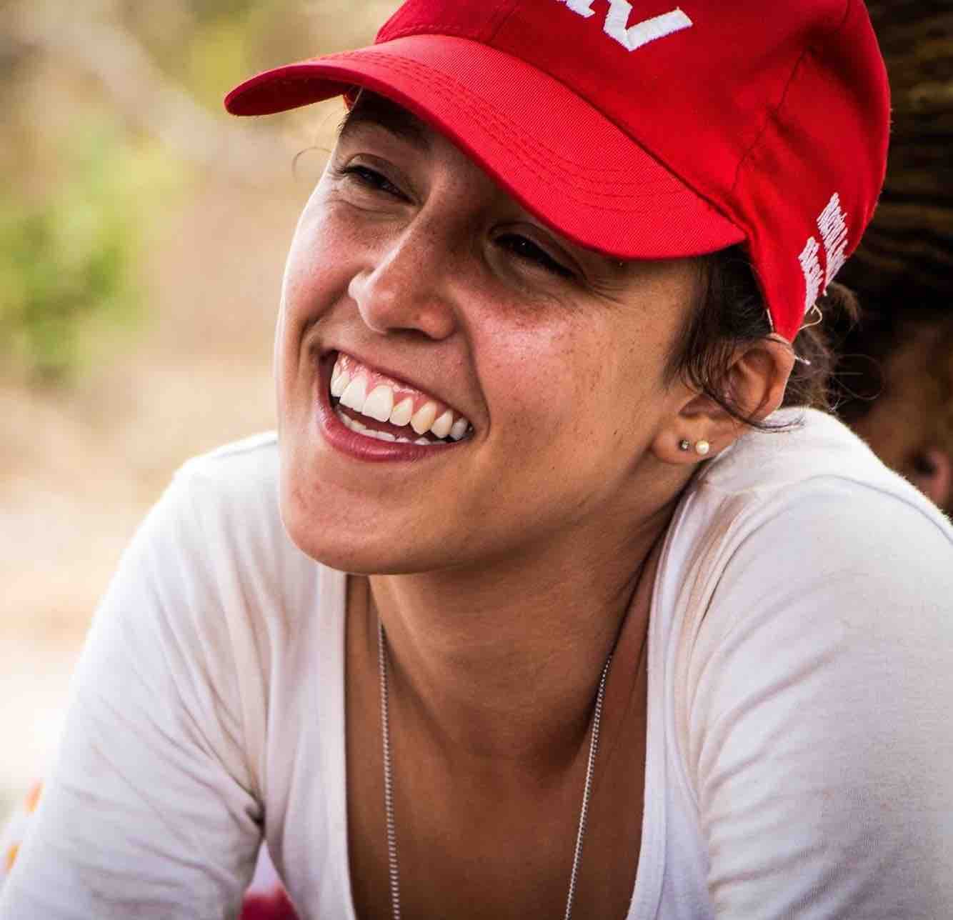 Ela é uma mulher branca, e na foto ela está com uma blusa branca, e seus cabelos castanhos aparecem pouco. Juliana usa um boné na cor vermelha, está posicionada levemente de lado e sorri olhando para a lateral. Ela usa um brinco pequeno perolado e uma corrente prateada.