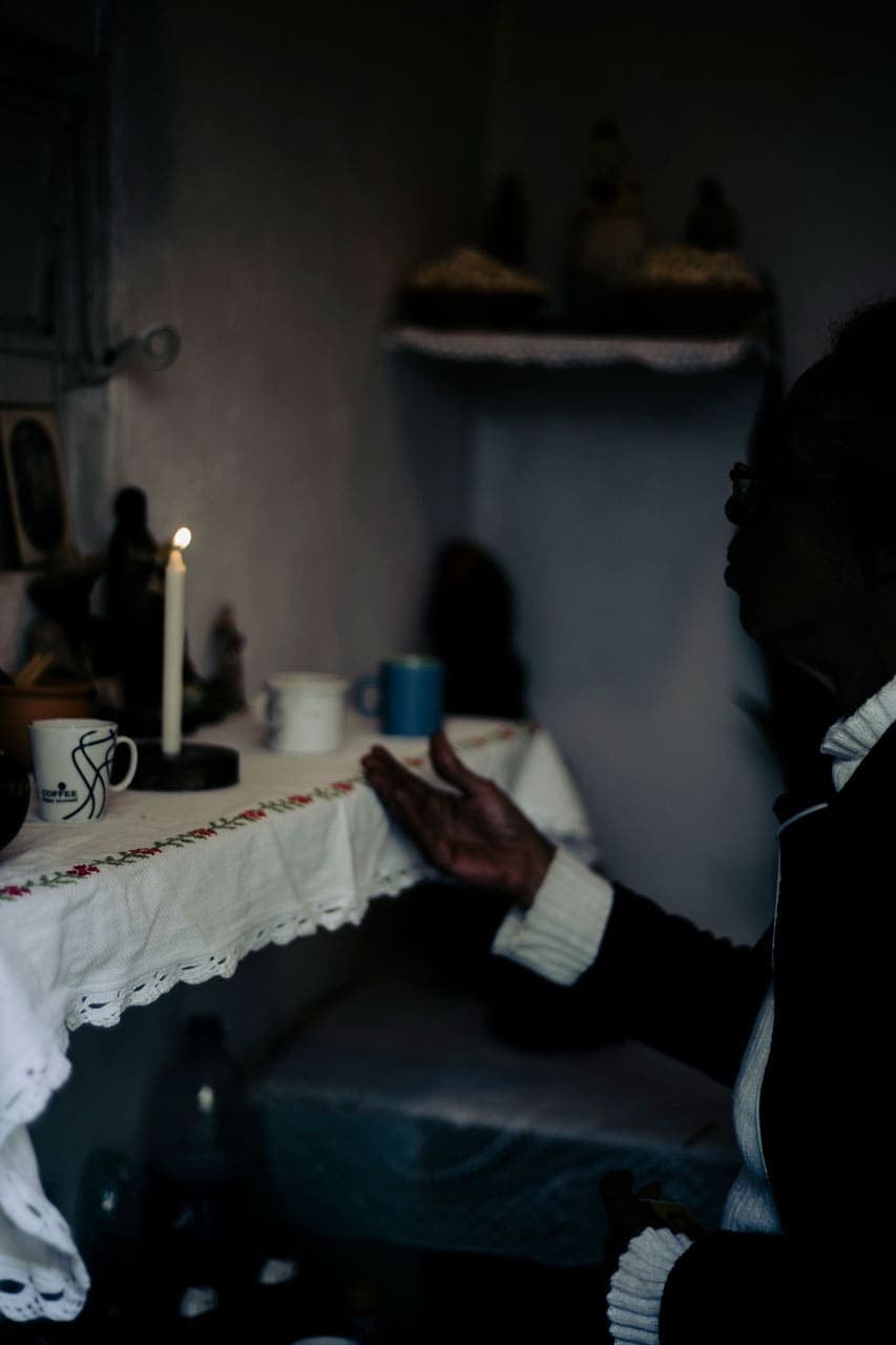 A foto traz a imagem de uma senhora em contraluz, ajoelhada em frente de um altar com uma das mãos estendidas. Neste altar, coberto por um pano com bordados de flor, algumas xícaras, imagens santas e uma vela acesa.