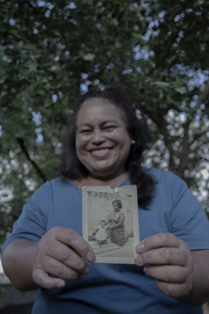 A foto mostra uma mulher em segundo plano, com blusa azul e cabelos parcialmente presos. Ela sorri pra câmera e mostra uma foto em preto em branco. A foto está em primeiro plano e mostra a imagem em preto e branco de uma mulher ajoelhada dando as mãos para um bebê com vestido que começa a andar.