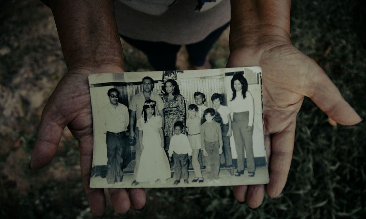 A foto mostra duas mãos abertas e estendidas segurando uma foto grande. Na foto, em preto e branco, 10 pessoas, sendo 4 adultos e 6 crianças. Todos usam roupas comuns, salvo uma das crianças que está ao centro, uma menina vestida com um longo vestido branco e uma coroa de flores na cabeça.