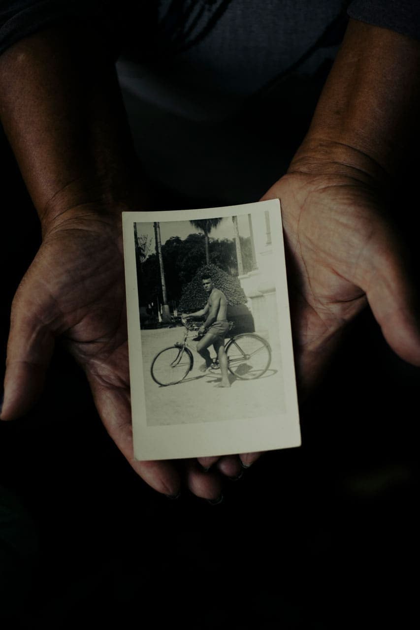 A foto mostra uma fotografia antiga sendo segurada por duas palmas de mãos abertas. Na fotografia antiga, é possível ver um homem de short curto encostado em sua bicicleta. Esse homem parece estar em uma praça devido às construções e árvores que vemos ao fundo da foto. A imagem da foto está em preto e branco.