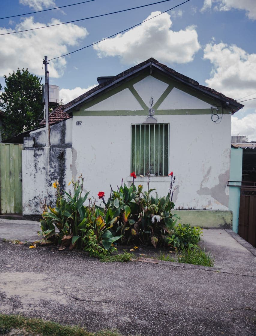 A foto mostra uma das casas da Cidade Ozanam de frente. O céu está azulado com algumas nuvens. A casa é branca e tem uma pintura desgastada. Alguns detalhes na empena e a janela estão pintados de verde e o telhado é de cerâmica em duas águas. Em frente a ela, cresce um pequeno canteiro de flores vermelhas e amarelas.