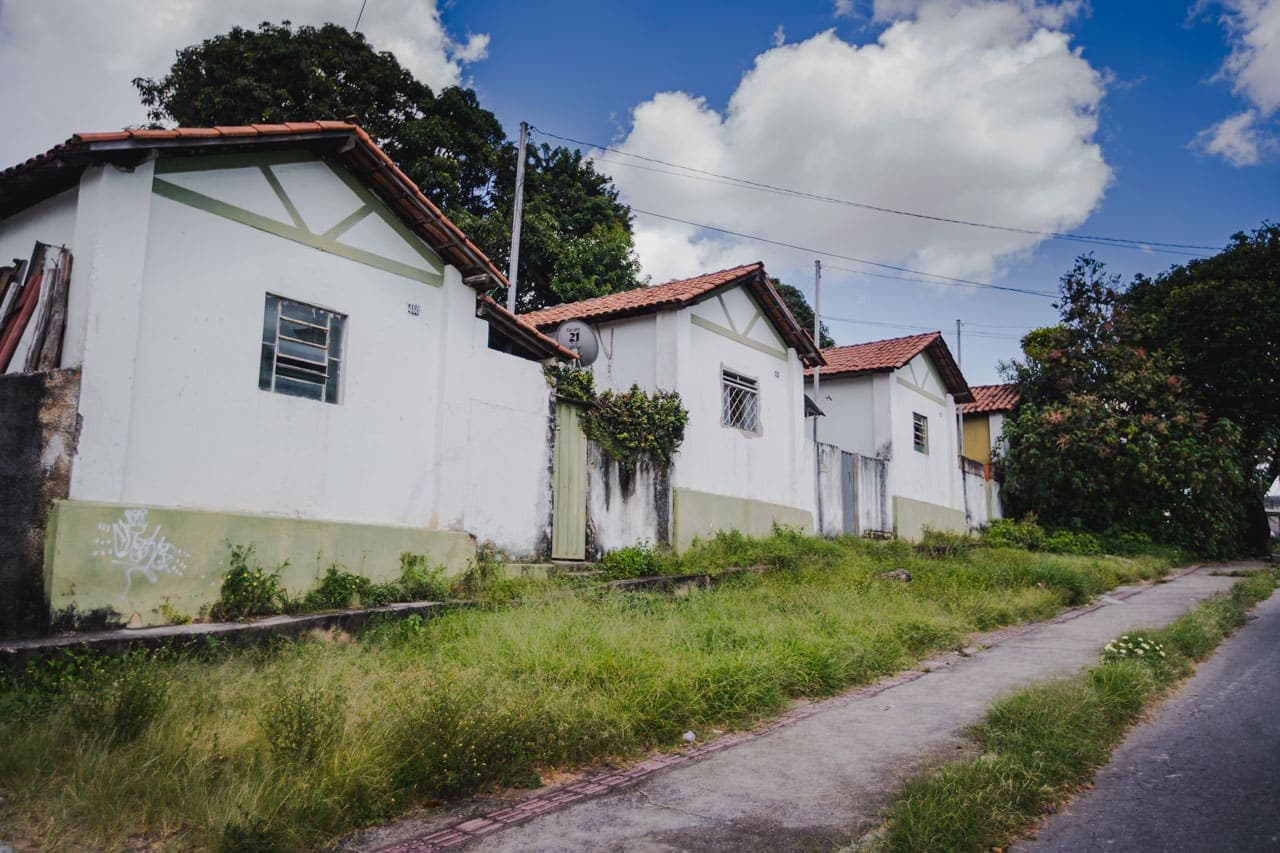 A foto mostra as casinhas enfileiradas do bairro Cidade Ozanam. As casas são brancas, com detalhes verdes e telhado de cerâmica. Em frente a elas, uma calçada gramada.