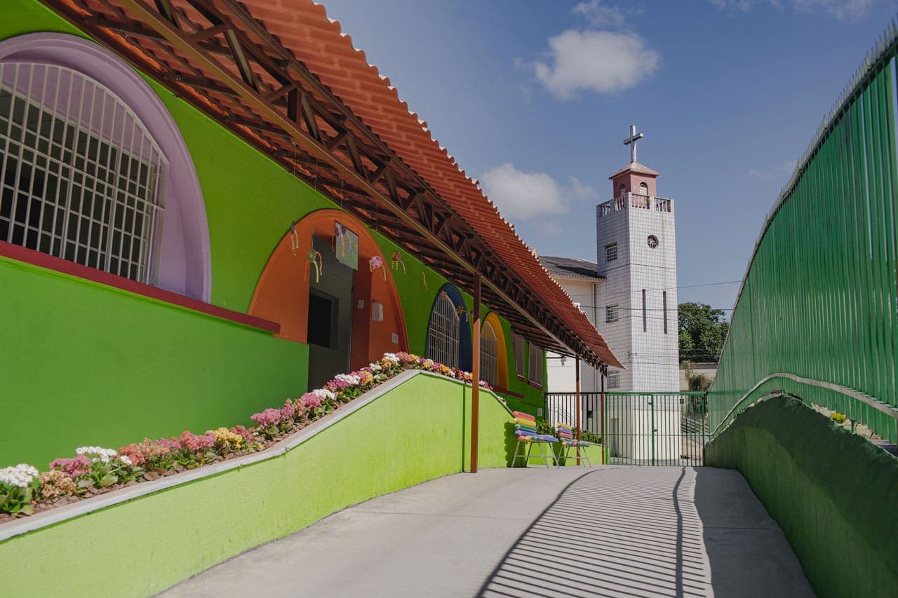 A foto mostra uma escola de telhado alaranjado, paredes verde limão, e portas e janelas emolduradas com tinta colorida. A rampa de acesso a escola, também verde limão, tem canteiros floridos. O chão é de cimento e a escola está cercada por grades verdes. Ao fundo, uma torre quadrada revestida com cerâmicas brancas. Na ponta da torre, uma pequena construção rosa com uma cruz no teto.