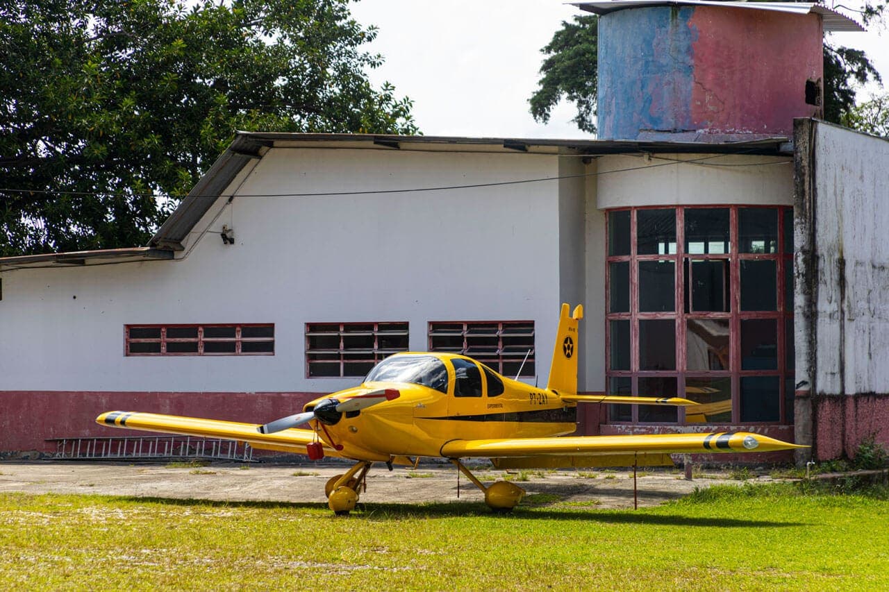 A foto mostra uma construção pintada de branco e vermelha. A pintura está desgastada. O chão à frente deste edifício é de cimento e, em seguida, uma grama verde. Estacionado entre o cimento e a grama, um avião de pequeno porte amarelo com detalhes em azul.