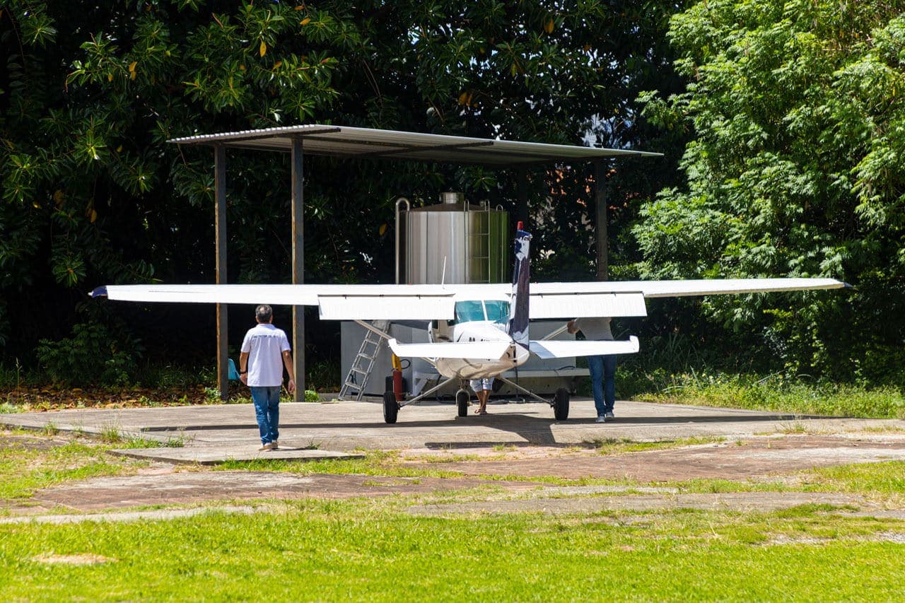 A foto traz mostra uma espécie de tanque de alumínio com cerca de 5 metros de altura, resguardado do sol por uma telhado feito de colunas de madeira e telha de amianto. A frente deste tanque está um avião de pequeno porte de cor branca e detalhes azuis. Um homem de blusa social e calça jeans caminha de costas para a imagem, em direção ao tanque. O tanque e o avião estão em um piso de cimento, rodeado por grama.