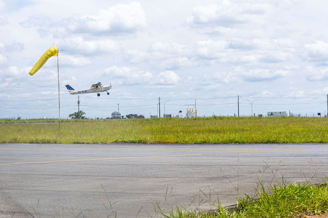 Nela, uma pista de asfalto cercada por grama e no horizonte um céu azul. À esquerda aparece um avião pequeno alçando voo. Mais à esquerda, uma biruta amarela indica a direção do vento.