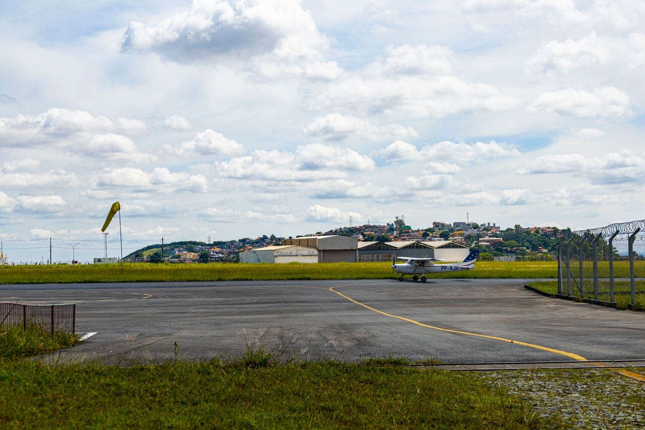 A imagem mostra a pista de pouso do Aeroporto Carlos Prates. A pista é rodeada por grama à frente e ao fundo. Na grama do fundo, alguns dos hangares onde as aeronaves são guardadas. Ao fundo da foto, já é possível ver parte do bairro Padre Eustáquio. Uma aeronave de pequeno porte branco com detalhes em azul está na pista.