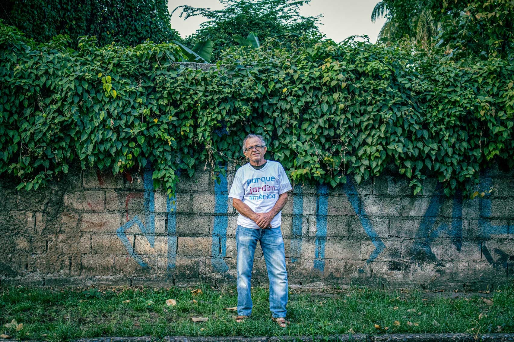 A imagem traz um senhor de sandália, calça jeans e blusa branca escrita “Parque Jardim América”. Ele tem os cabelos brancos, usa óculos e está com as mãos à frente do corpo. Ele está em frente ao muro do terreno onde será o Parque. O muro é cinza e as árvores e plantas encobrem o final do muro.