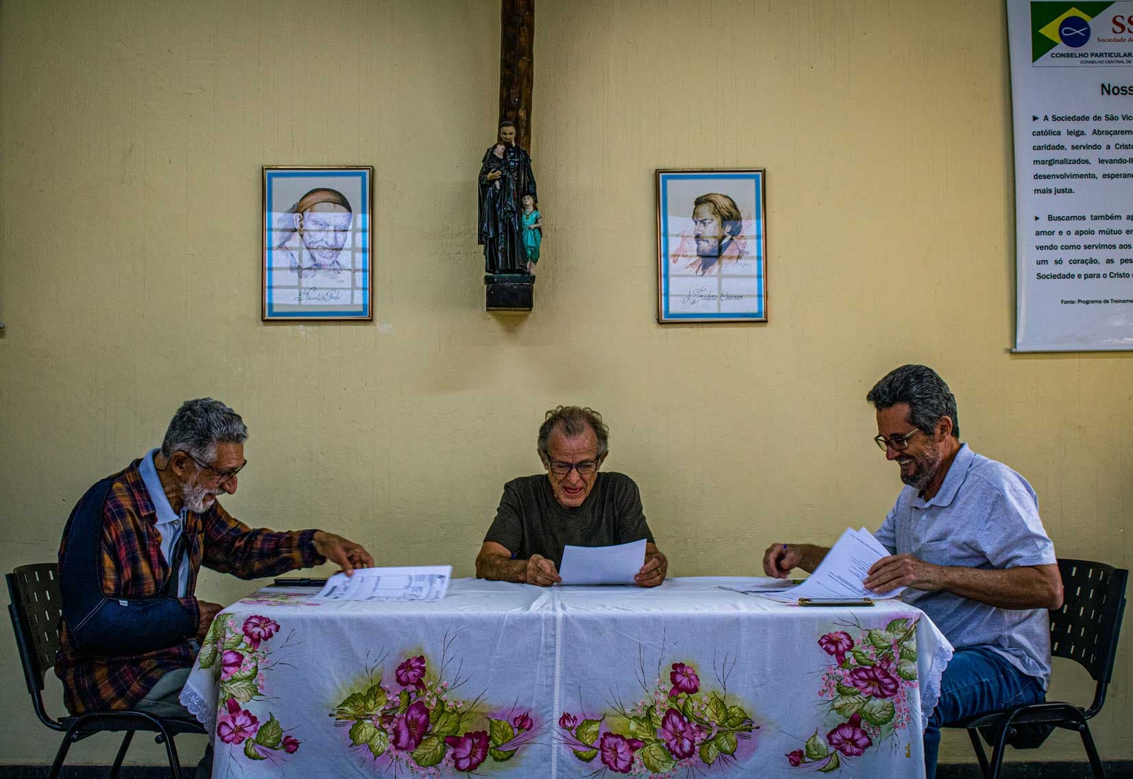 A foto mostra três homens sentados em uma mesa observando papéis. Os três homens têm cabelos grisalhos e usam óculos. As cadeiras em que estão sentadas são pretas e a mesa está coberta por um pano branco com pinturas de flores rosas e folhas verdes. Ao centro, uma imagem de um santo perto de duas crianças. Na parede de cor creme, um quadro de cada lado da imagem do santo.