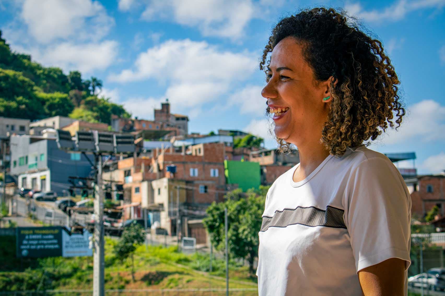 A foto traz a imagem de uma mulher de cabelos de tamanho médio, enrolados, olhando para a esquerda e sorrindo. Ela usa uma camisa branca com uma listra cinza na altura das mangas. Ao fundo, um espaço verde e casas de tijolos aparentes.