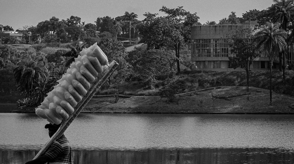 A imagem mostra uma paisagem com uma lagoa abaixo e uma construção moderna do lado direito. O restante são árvores e vegetação ao redor. Do lado esquerdo da foto, aparece um vendedor de algodão doce com os sacos pendurados em um pau que ele ergue com as mãos.