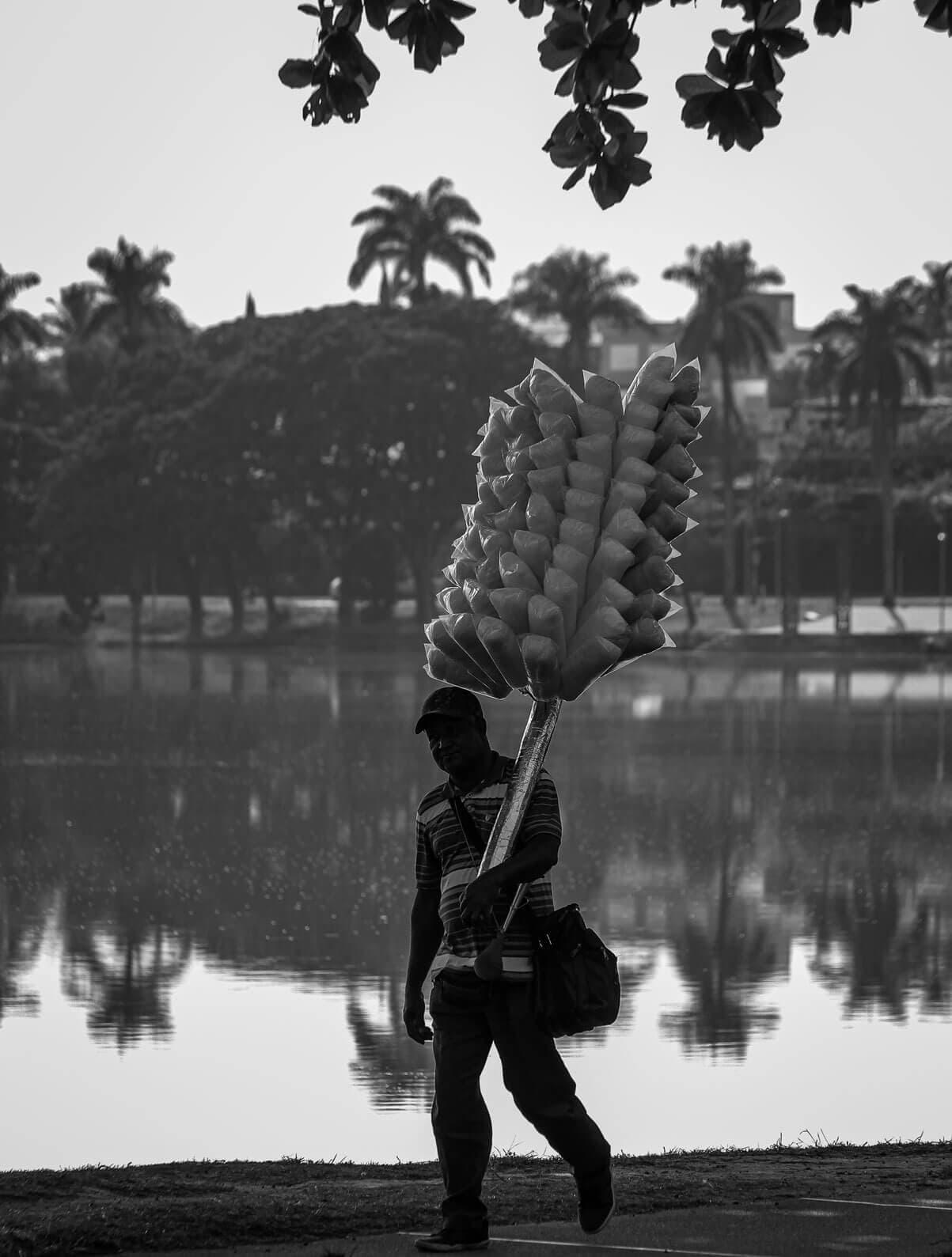 A imagem tem, ao fundo, a Lagoa da Pampulha, rodeada de árvores. No primeiro plano, temos um homem vendendo algodão doce, com uma bolsa de lado e olhando para o fotógrafo.