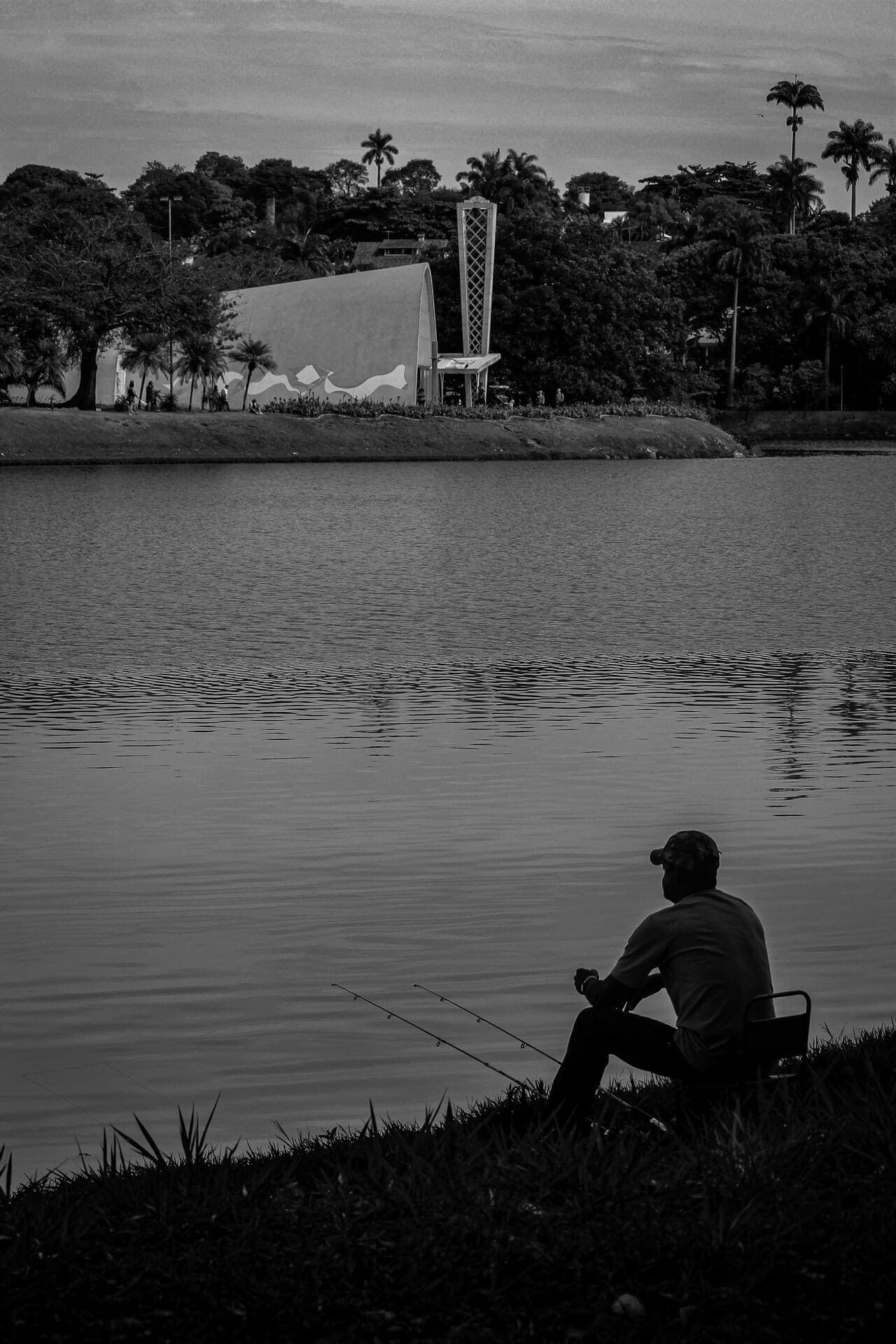 A imagem é ocupada pela Lagoa da Pampulha, em sua maior parte. No fundo, é possível ver a Igreja São Francisco de Assis. No primeiro plano da foto, vemos um homem sentado na grama observando a paisagem.