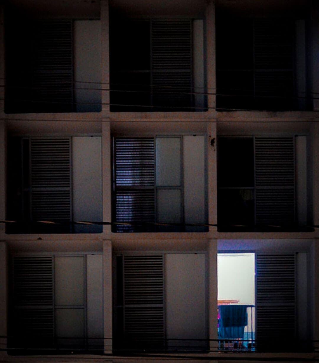 A imagem mostra um conjunto de janelas/varandas de um prédio. A foto está escura e parece ter sido tirada de noite. Em um dos apartamentos, é possível ver uma luz acesa e a porta aberta. Tem uma toalha estendida na grade da varanda.