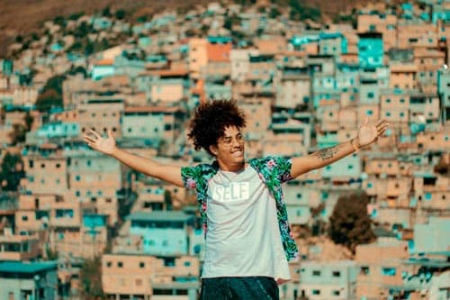 Rafael Freire é um homem jovem, de pele negra e cabelos black power. Aparece de braços abertos, vestindo uma camisa colorida estampada por cima de uma blusa de cor clara, Ao fundo, vê-se a paisagem do Aglomerado da Serra.