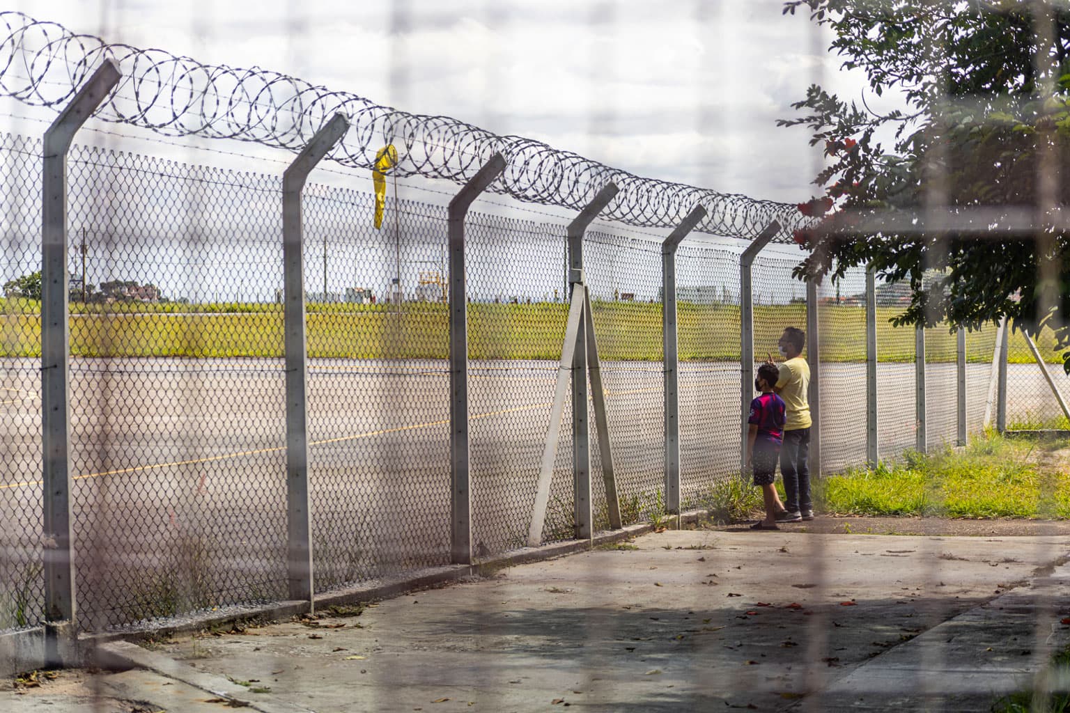 A foto mostra um homem e uma criança observando o Aeroporto Carlos Prates do lado de fora, por entre os buracos de uma tela de proteção que separa o aeroporto do restante da rua. O homem usa blusa amarela, calça preta e tênis. A criança usa uma blusa azul e vinho, bermuda preta e chinelo.