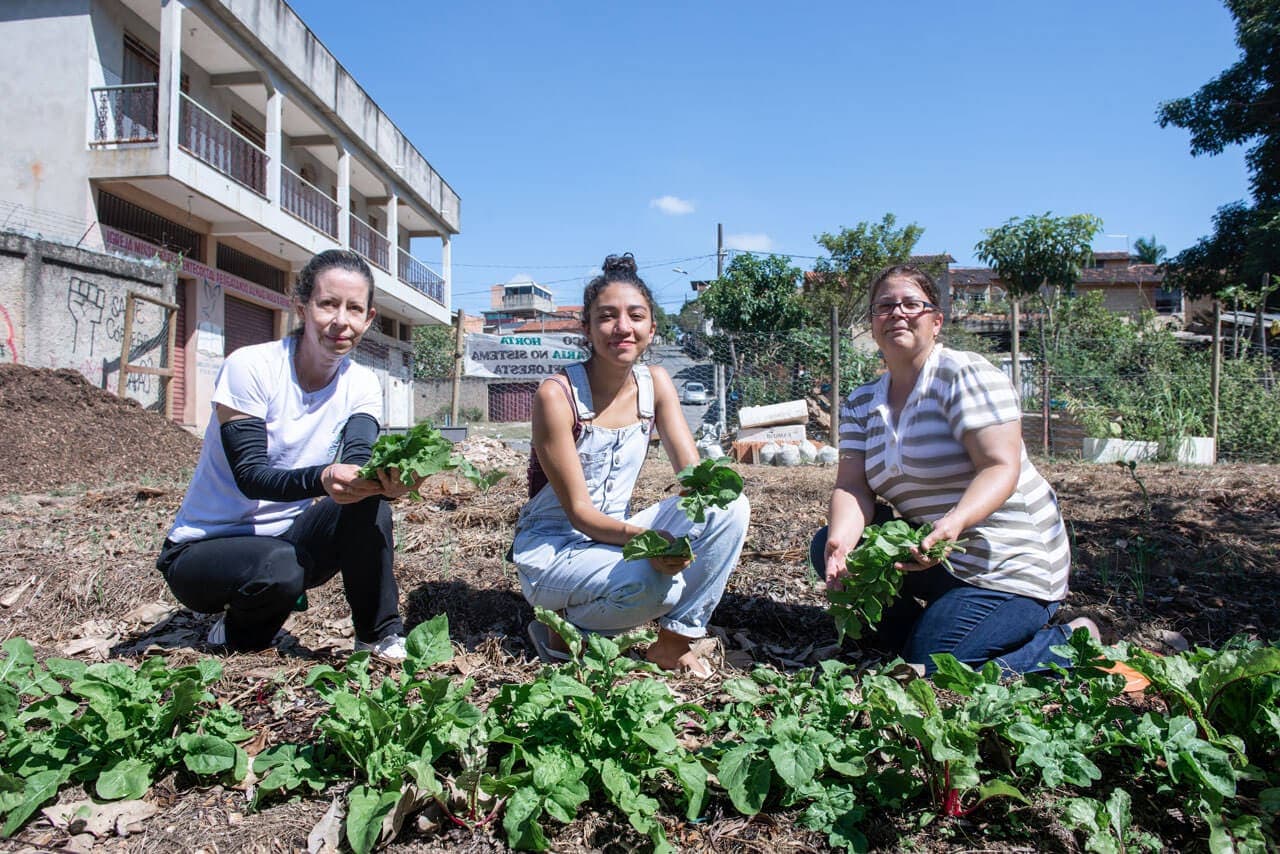 A imagem reúne, em primeiro plano, três mulheres sentadas ao chão, em frente a um pequeno canteiro de hortaliças. Ao centro, a mais jovem das três, de pele parda e cabelos presos, sorri de olhos cerrados diante do sol quase a pino. A seu lado, há duas mulheres de cabelos igualmente presos e pele clara. Todas ostentam verduras recém-colhidas.